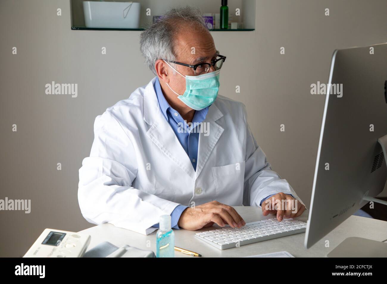Medico di sesso maschile senior che indossa maschera facciale e abito medico seduto al tavolo e parlare con il paziente tramite videochiamata attiva Computer mentre lavora in remoto durante lo scoppio di COVID 19 Foto Stock