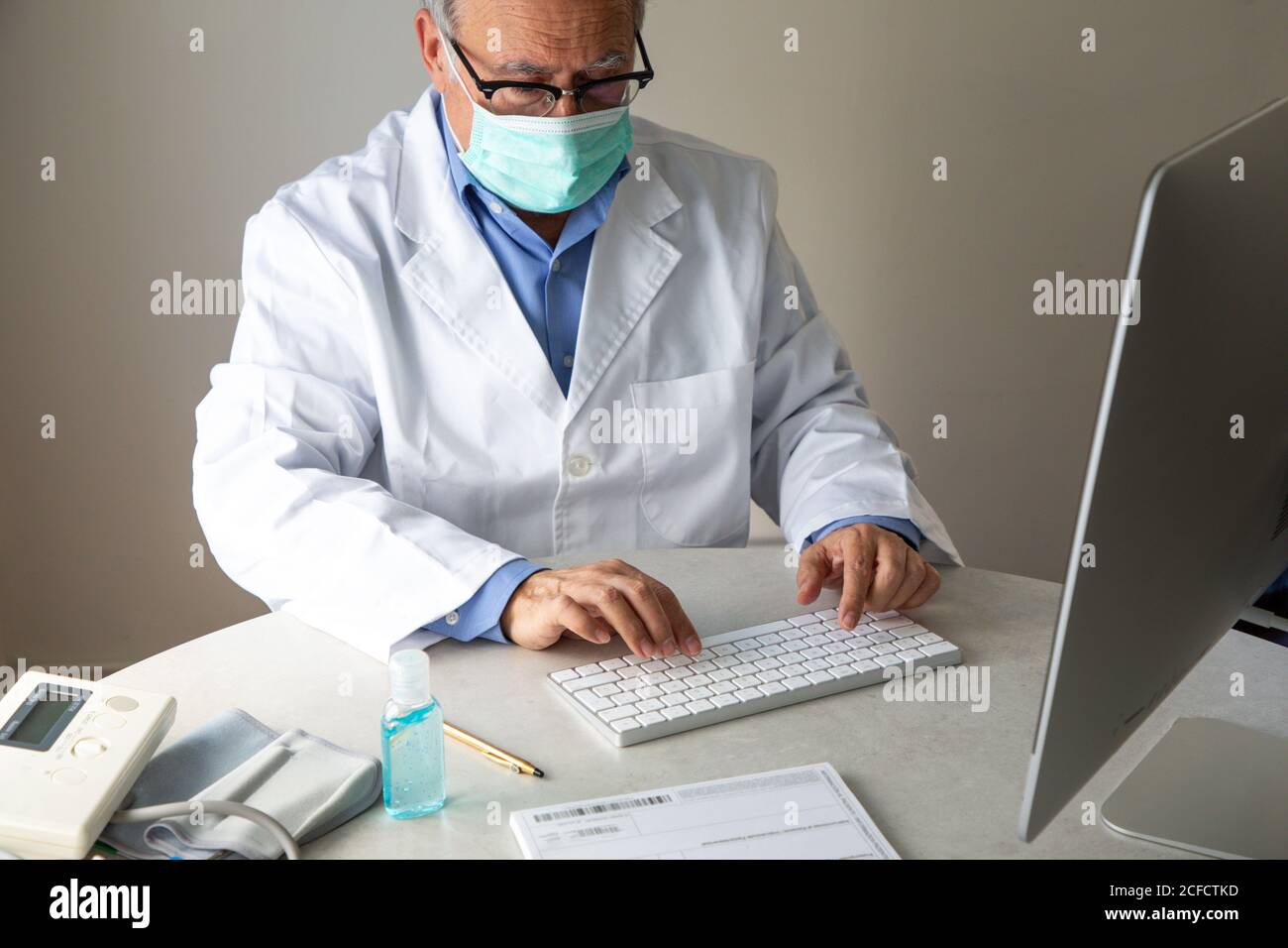Medico di sesso maschile senior che indossa maschera facciale e abito medico seduto al tavolo e parlare con il paziente tramite videochiamata attiva Computer mentre lavora in remoto durante lo scoppio di COVID 19 Foto Stock