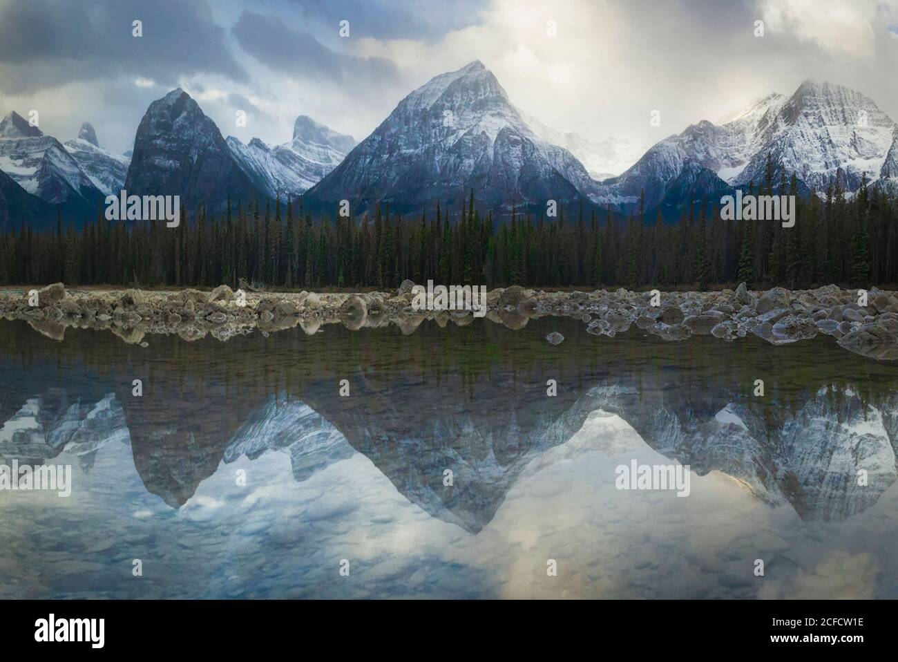 Scenario pittoresco con maestose montagne rocciose coperte di neve e. cielo nuvoloso riflesso in lago con foresta di conifere sulla riva Nella campagna canadese Foto Stock