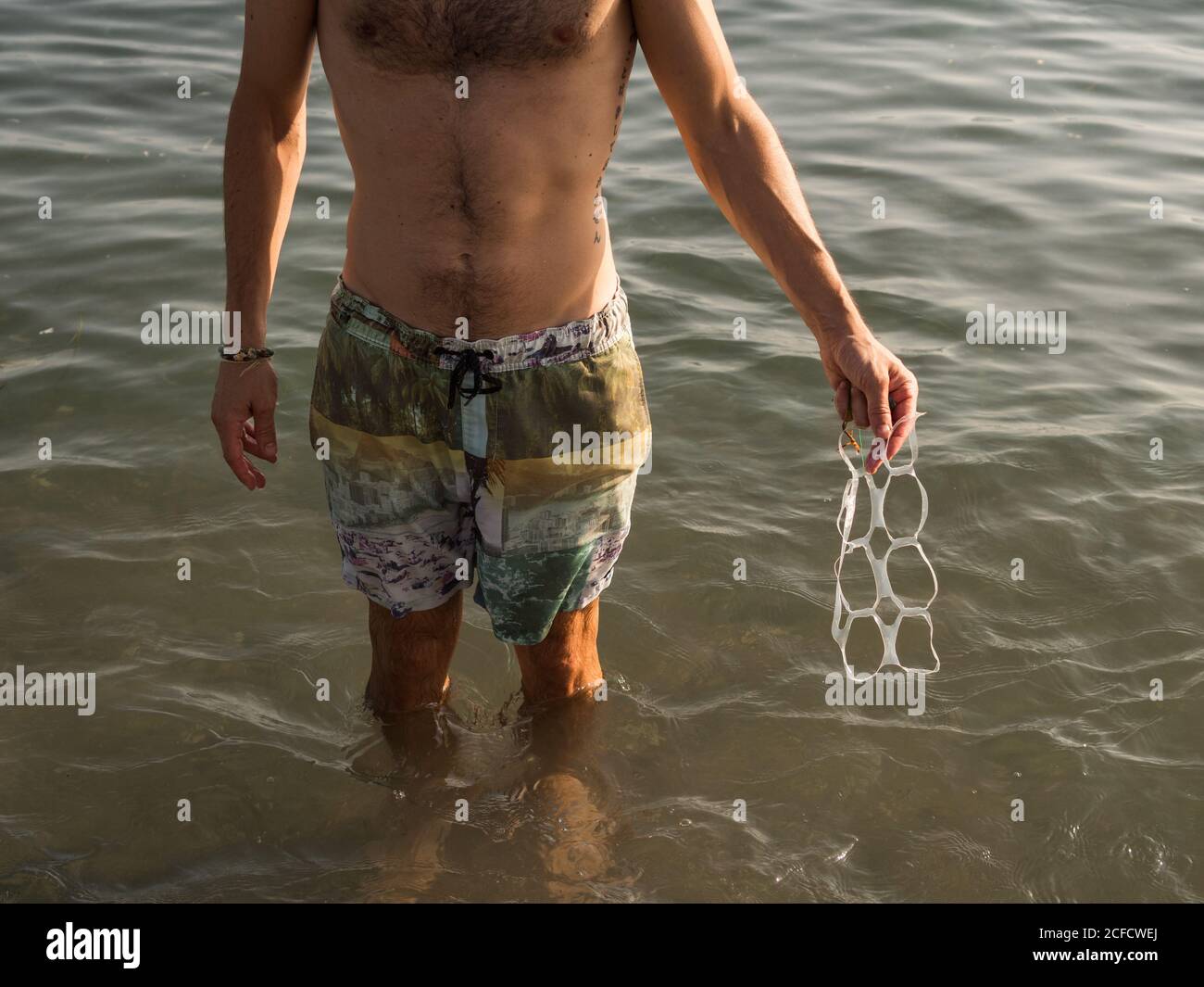Da sopra anonimo maschio con petto peloso che trasporta rifiuti di plastica mentre si è in piedi in acqua di mare sul resort Foto Stock