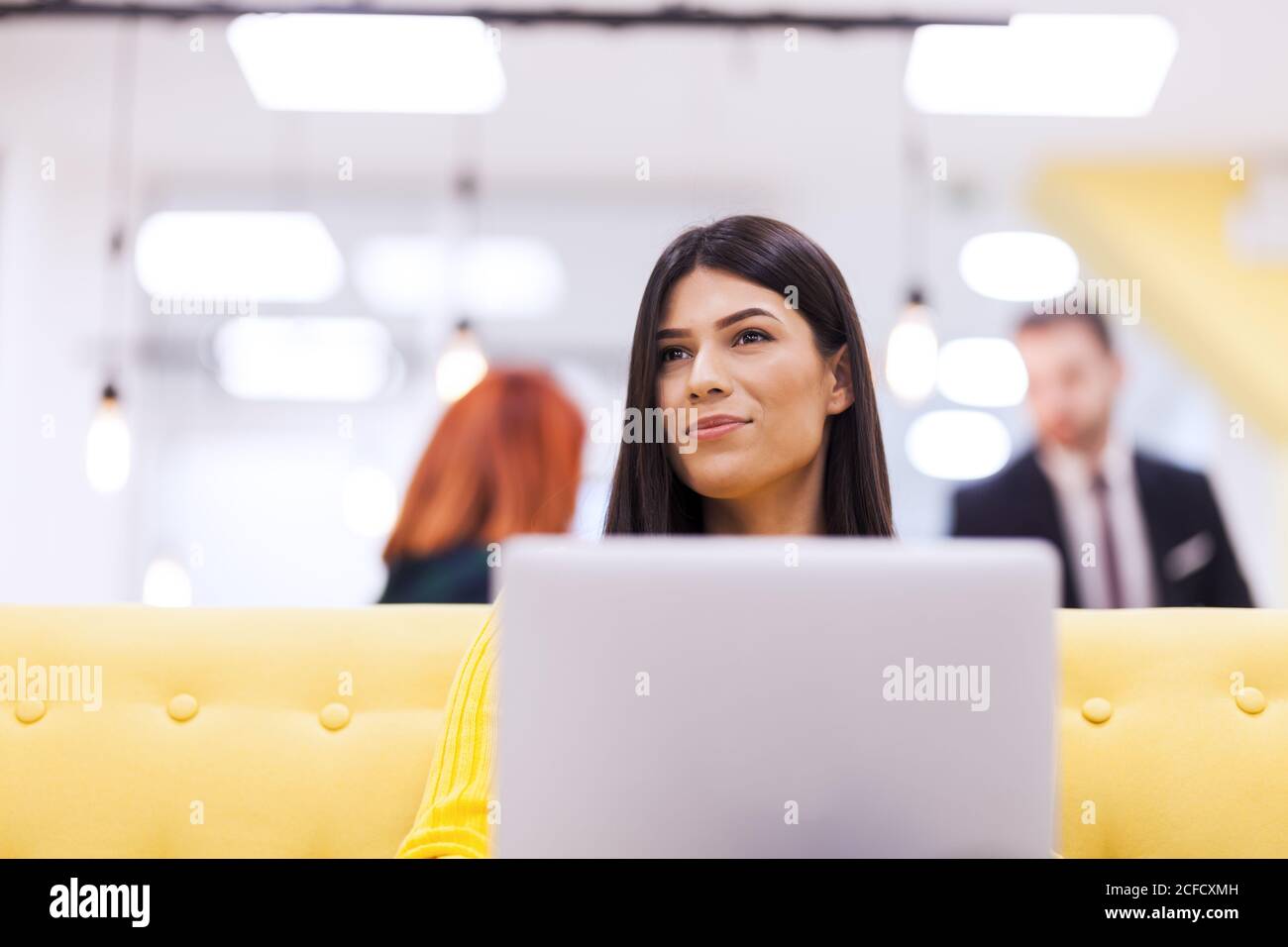 Bella ragazza con lunghi capelli scuri che lavora su un computer portatile in un ufficio moderno. Colleghi di affari nel backgorund Foto Stock