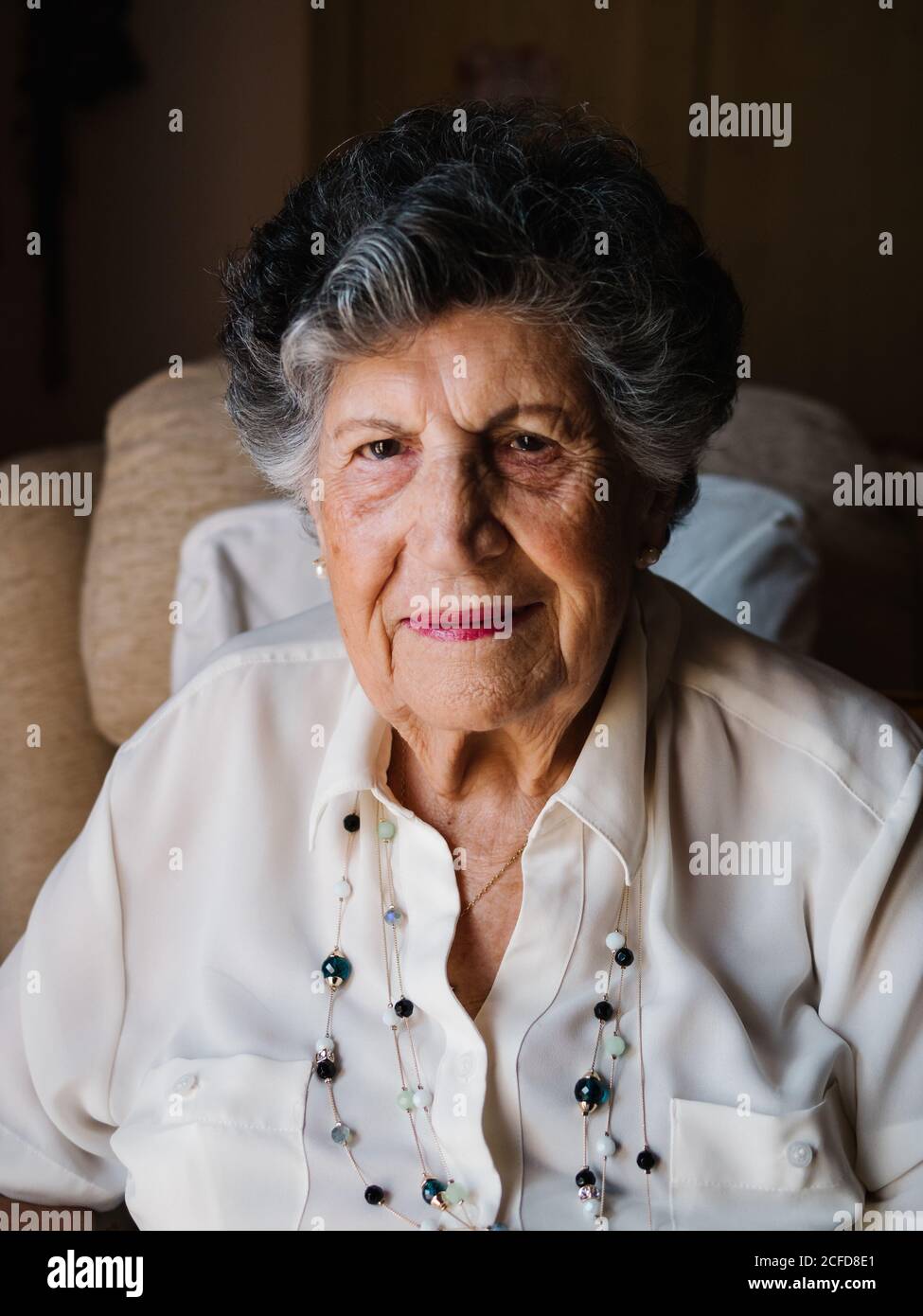 Ritratto di felice donna con capelli ricci grigio in camicia bianca e con perline sul collo guardando la macchina fotografica a casa Foto Stock