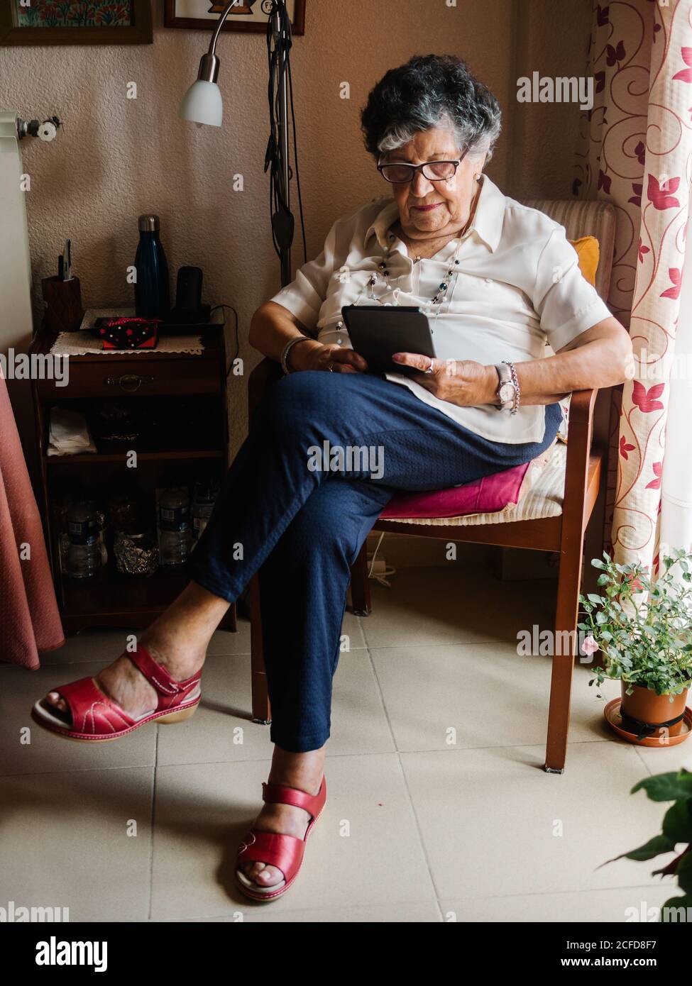 Donna anziana dai capelli grigi in camicia e bicchieri seduta su poltrona e lettura e-book in appartamento Foto Stock