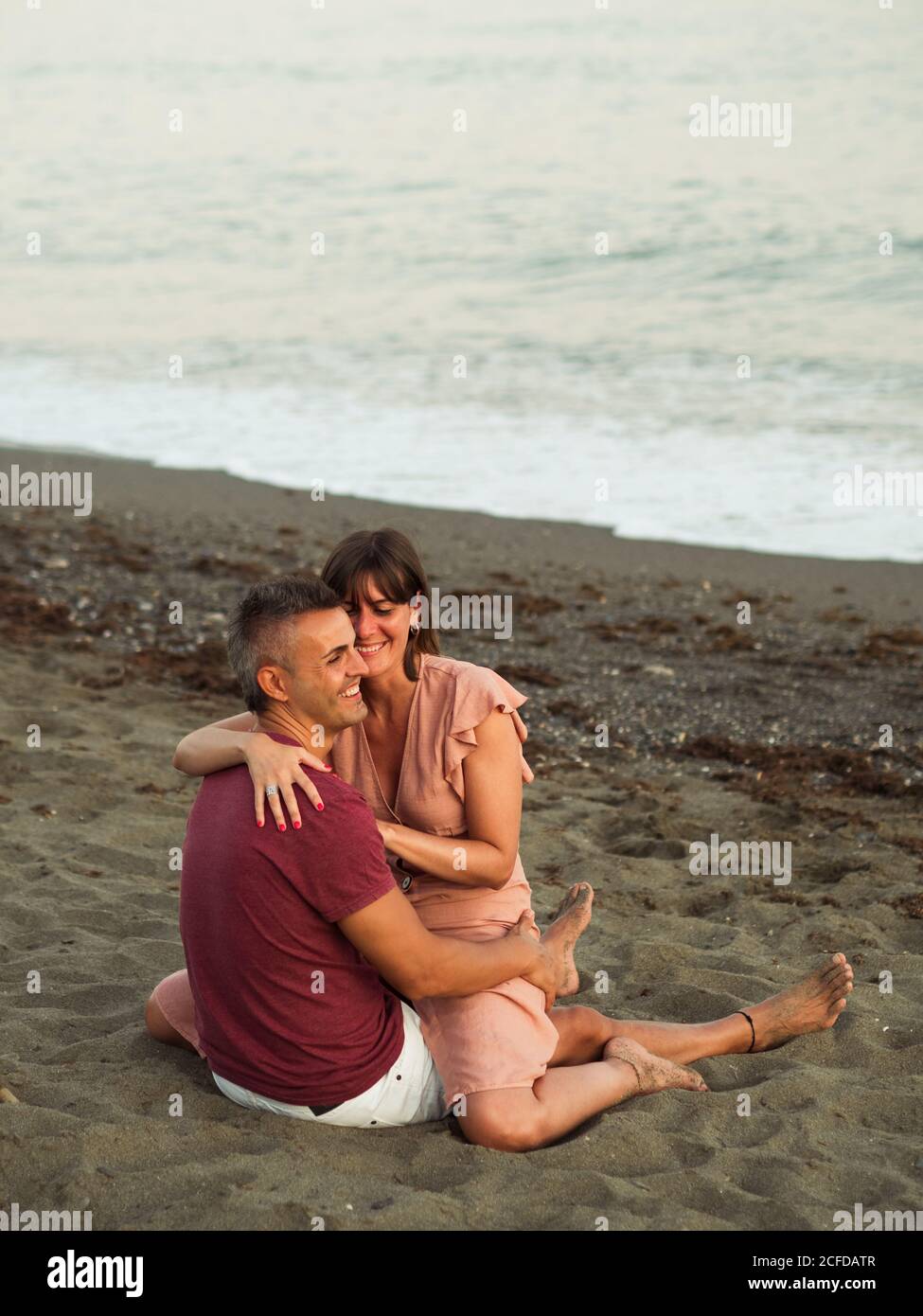 Uomo e donna adulta corpo pieno sorridente e abbracciante mentre si siede sulla sabbia vicino al mare e si rilassa durante la data Foto Stock