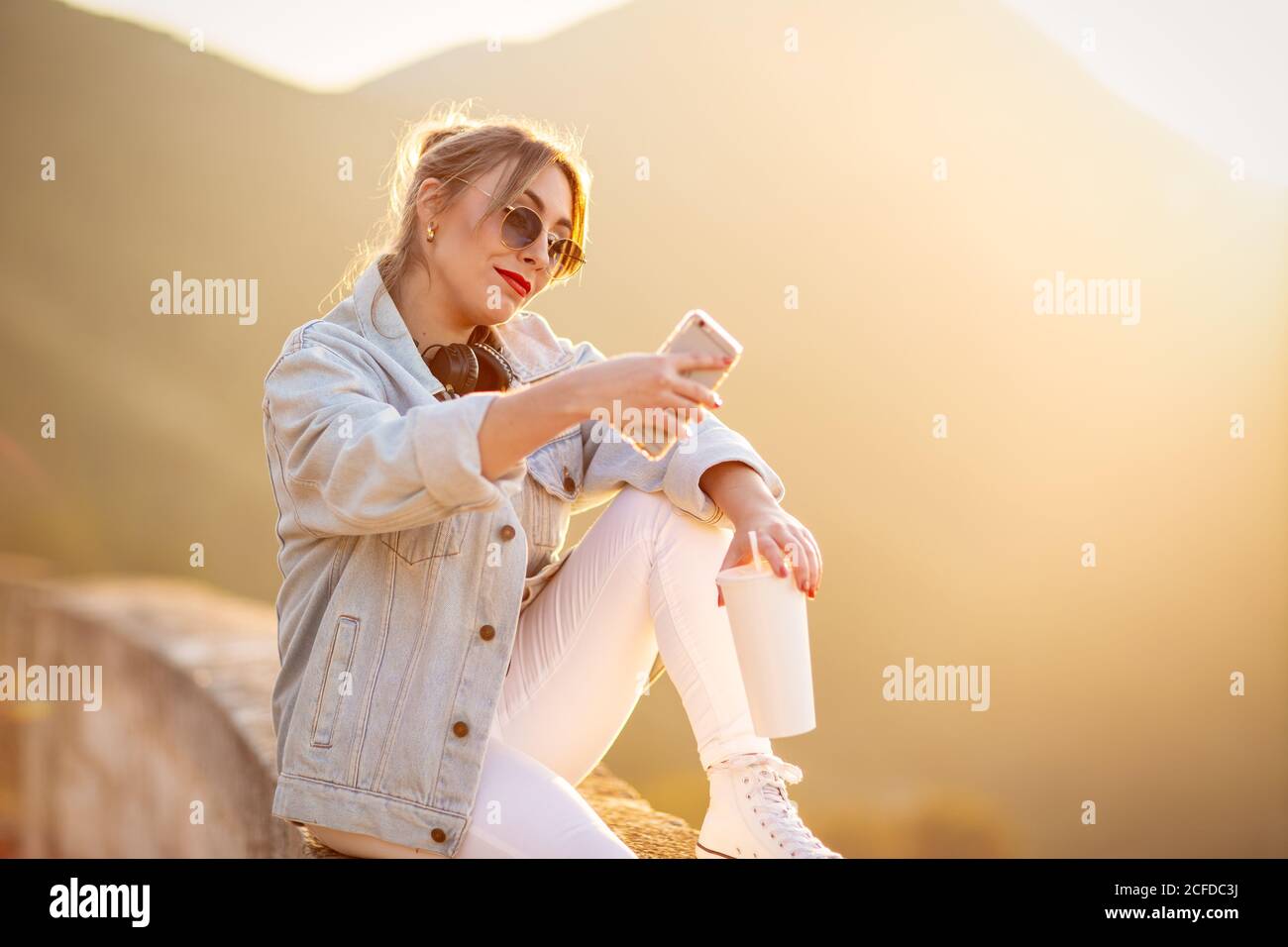 Signora carina in occhiali da sole alla moda sorridente mentre prendendo selfie sopra telefono cellulare con luce calda del sole su sfondo sfocato Foto Stock