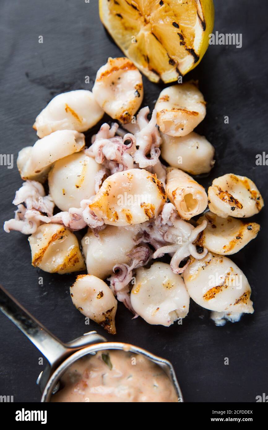 Un mucchio di deliziosi calamari alla griglia e piccoli polpi posti vicino limone e salsa al tavolo nel caffè Foto Stock