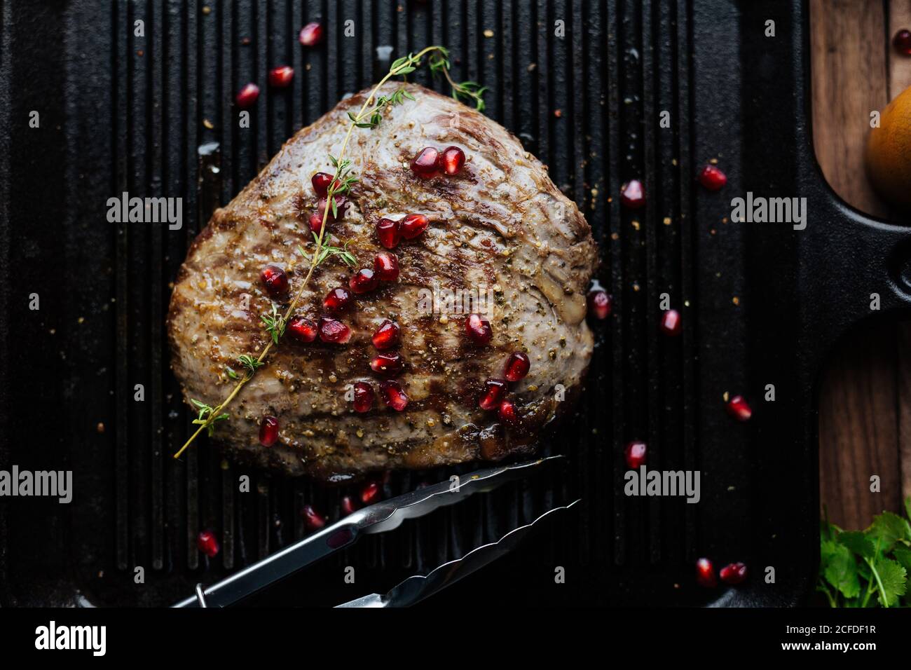 Vista dall'alto della bistecca di manzo appetitosa guarnita con semi di melograno ed erbe servite su piastra nera con le pinze della cucina sopra tavolo di legno Foto Stock