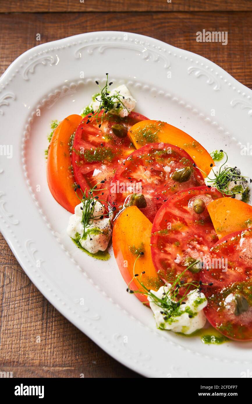 Dall'alto di fette rosse fresche di pomodori e mango su piatto bianco decorato con erbe feta e pesto salsa Foto Stock