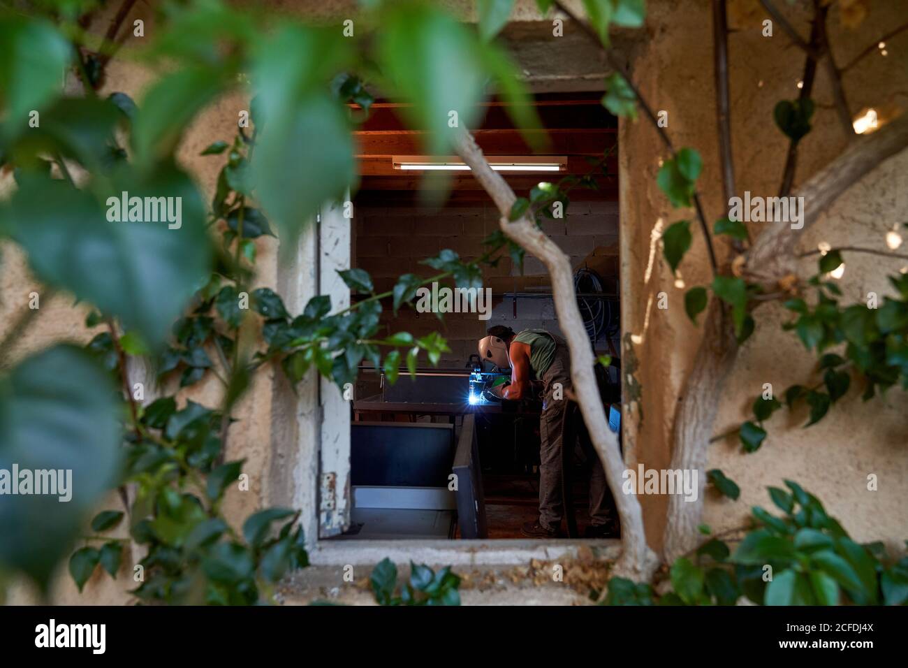 Viste da fronde di rami di albero del maestro professionista in protezione maschera di saldatura metallo mentre si lavora in piccole officine Foto Stock