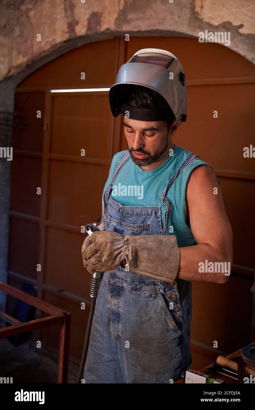 Uomo professionista con maschera di saldatura sulla testa indossando tute in jeans e guanti di protezione in piedi sul luogo di lavoro e preparazione dello strumento per saldatura Foto Stock