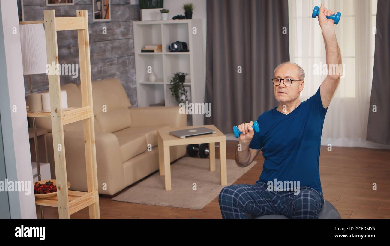 Uomo anziano che fa la pressa della spalla con i manubri nel soggiorno. Anziano pensionato sano addestrando sport di salute a casa, esercitandosi attività di idoneità all'età anziana Foto Stock