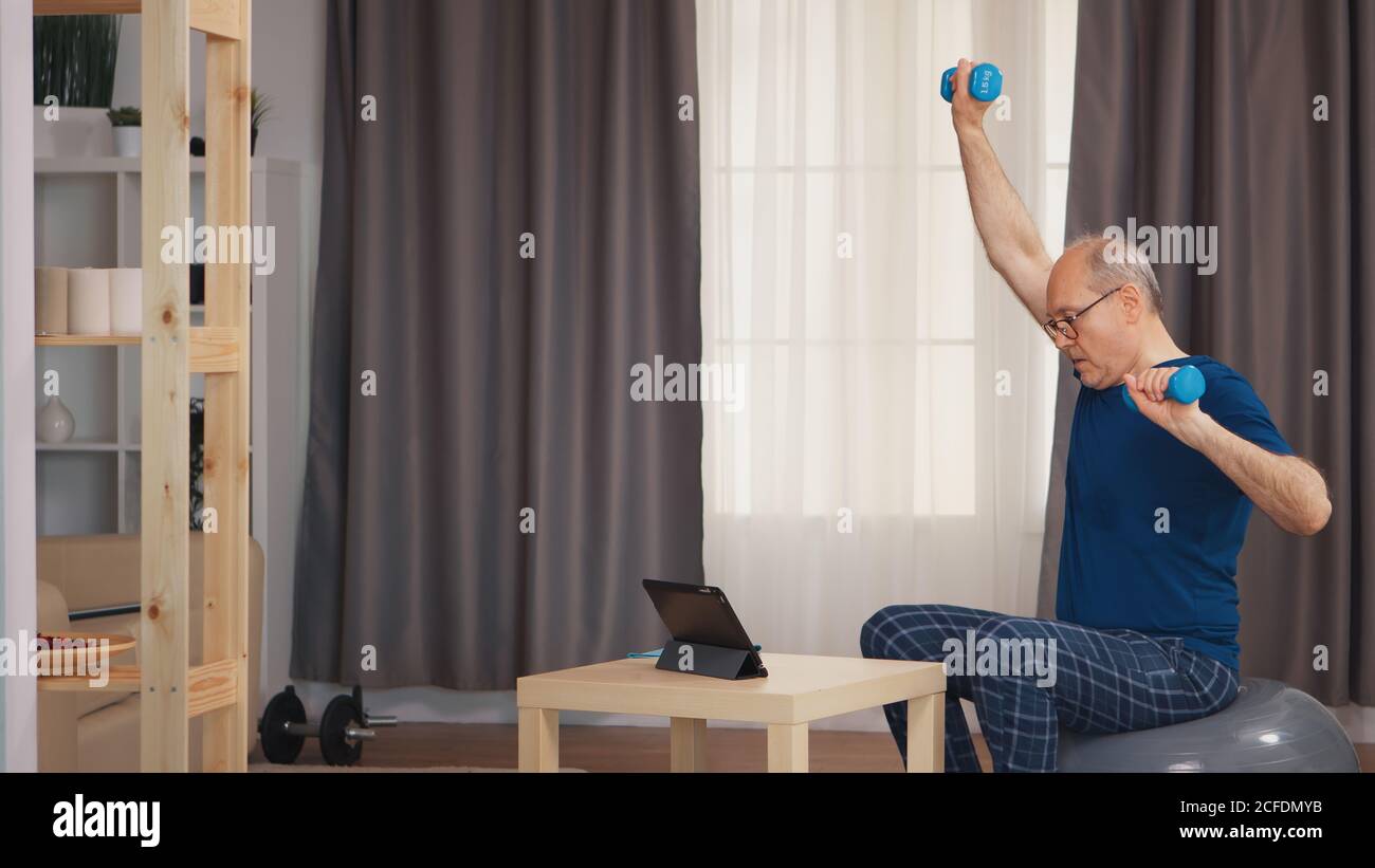 Allenamento nonno in soggiorno con manubri che guardano il programma di fitness online. Anziano pensionato sano addestrando sport di salute a casa, esercitandosi attività di idoneità all'età anziana Foto Stock