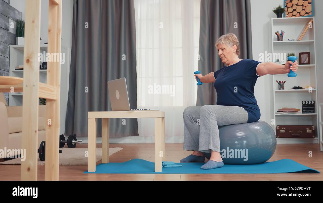 Donna anziana sana che si esercita con manubri in soggiorno. Allenamento online in internet per anziani a casa sport stile di vita sano, allenamento fitness per anziani in appartamento, attività e assistenza sanitaria Foto Stock