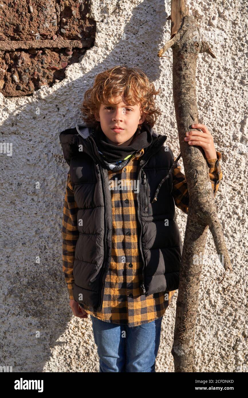 Carino bambino curly in gilet turistico caldo in piedi vicino al muro e tenendo asciutto tronco di albero in giorno luminoso Foto Stock