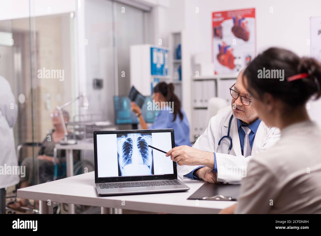 Medico maturo che punta alla scansione tc dei polmoni del paziente sul computer portatile durante la visita medica in ospedale. Medico in camice bianco che discute con la donna anziana disabile in sedia a rotelle e infermiera nella presa dei raggi X. Foto Stock