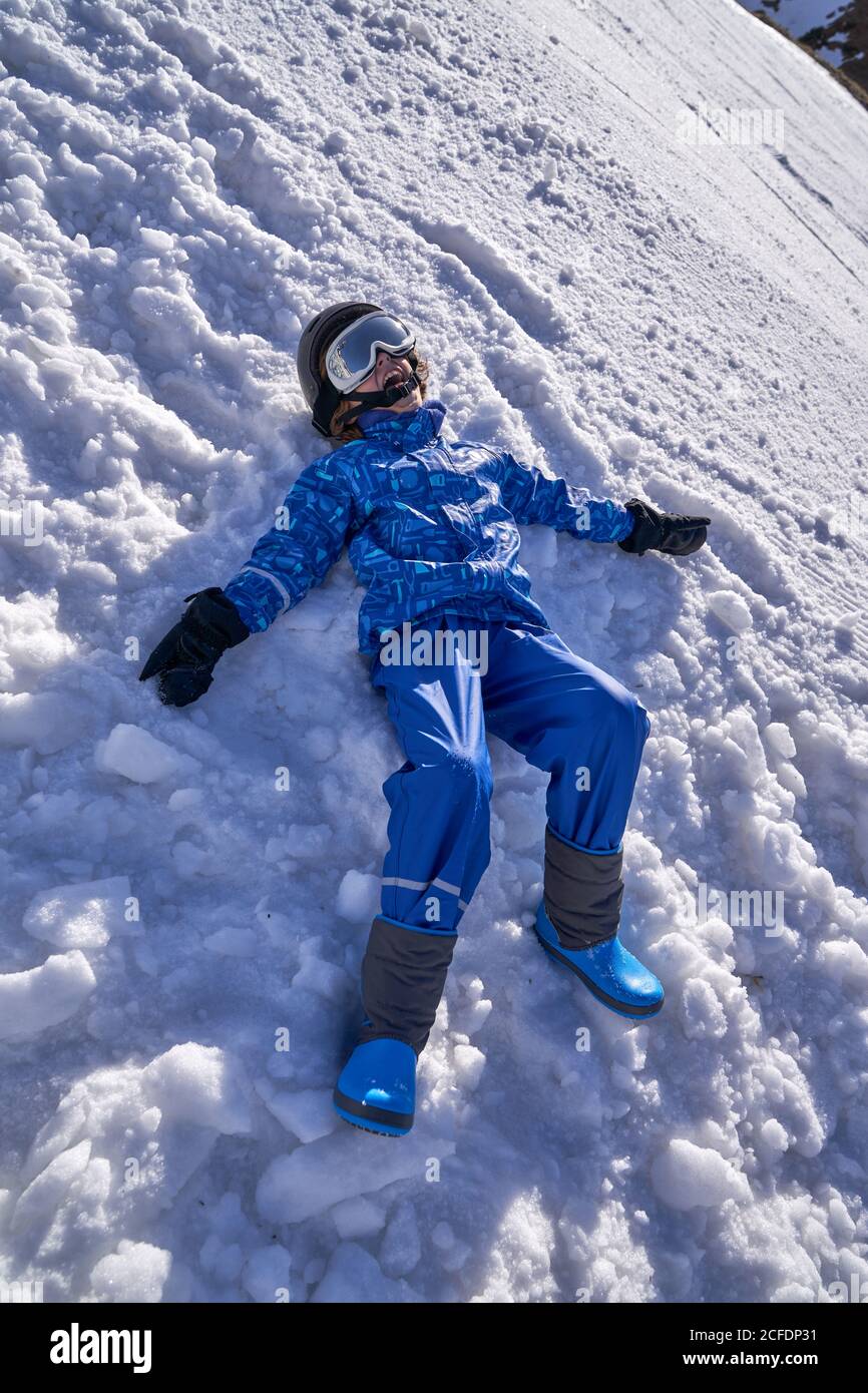 Ragazzo in abiti sportivi invernali e casco da snowboard con occhiali di  protezione giacenti su un terreno coperto di neve e iniziando a fare angelo  di neve mentre si ride felicemente a
