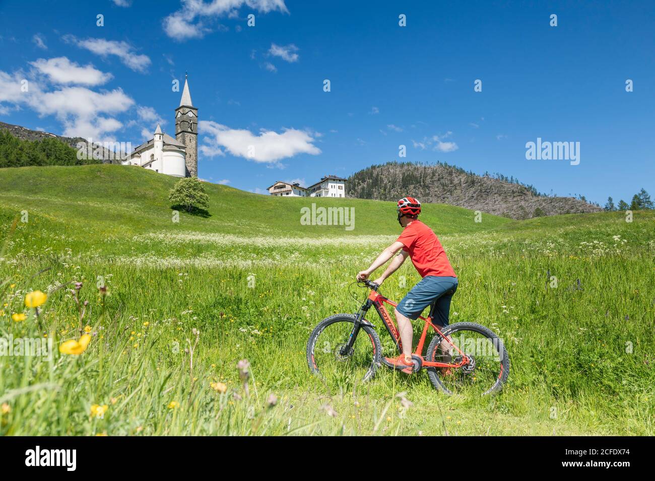 ciclista in e-bike a laste di rocca pietore, nei prati di fronte alla chiesa parrocchiale, dolomiti, agordino, belluno, veneto, italia Foto Stock