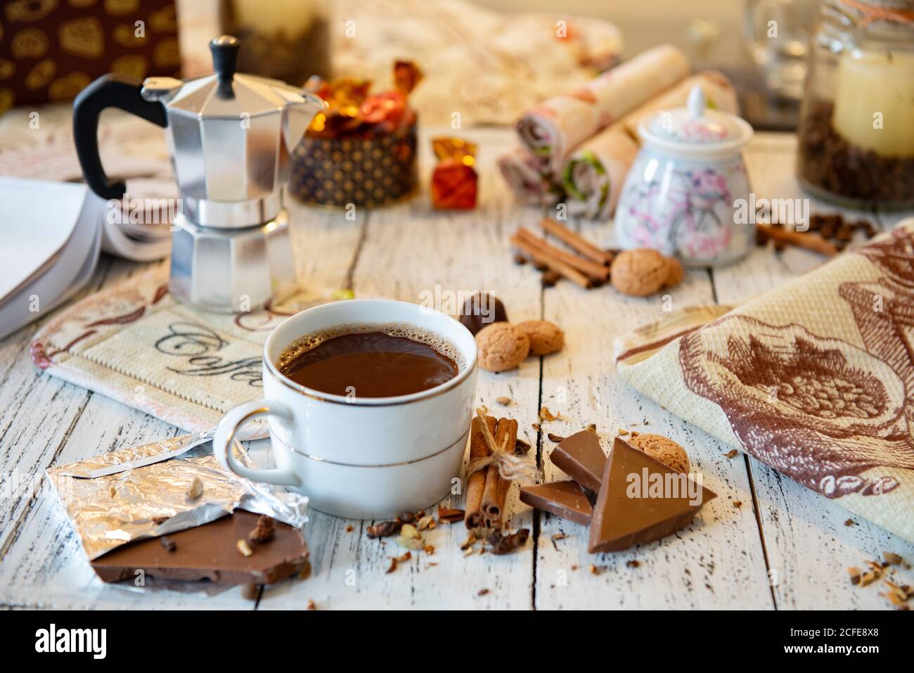 Tazza di caffè, cioccolato, chicchi di caffè, bastoncini di cannella e spezie su un tavolo di legno bianco. Buona colazione del mattino o pausa caffè sullo sfondo. Foto Stock