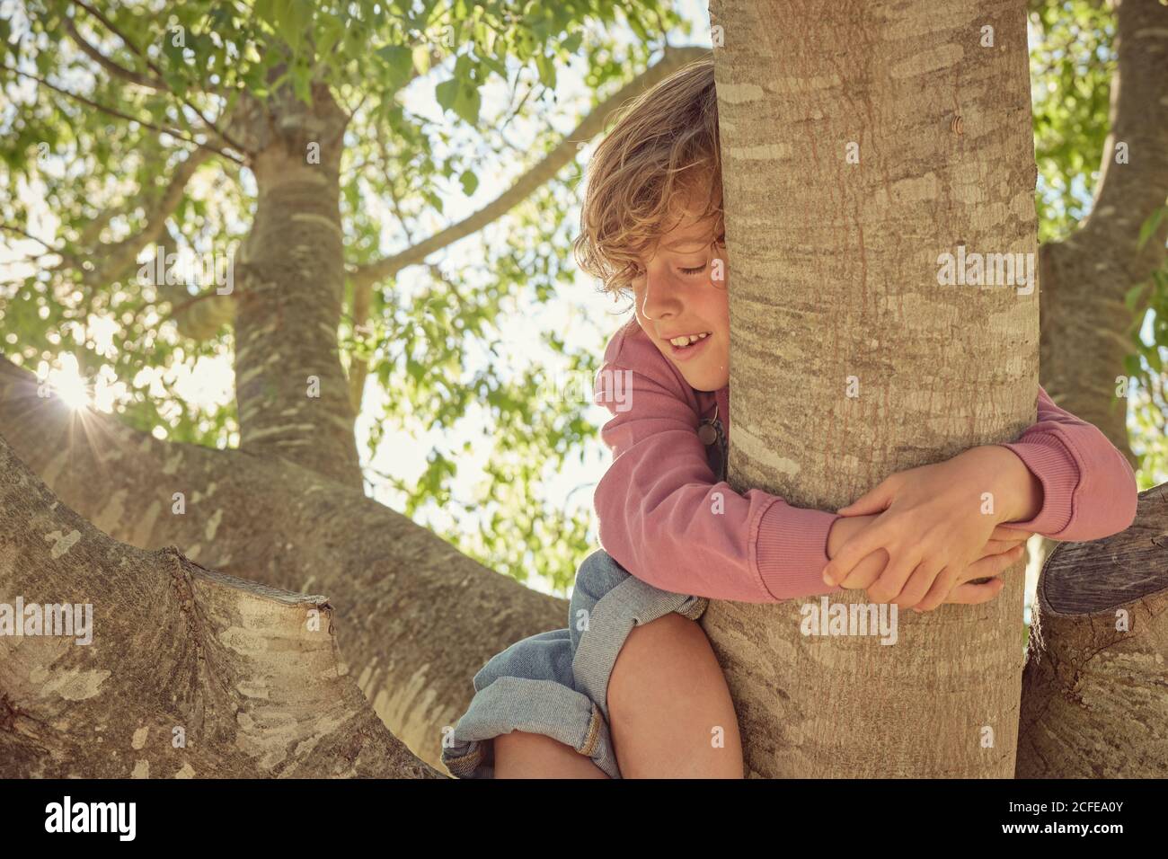 Sorridente bambino energico con gli occhi chiuso arrampicata albero e tenere su al trunk Foto Stock