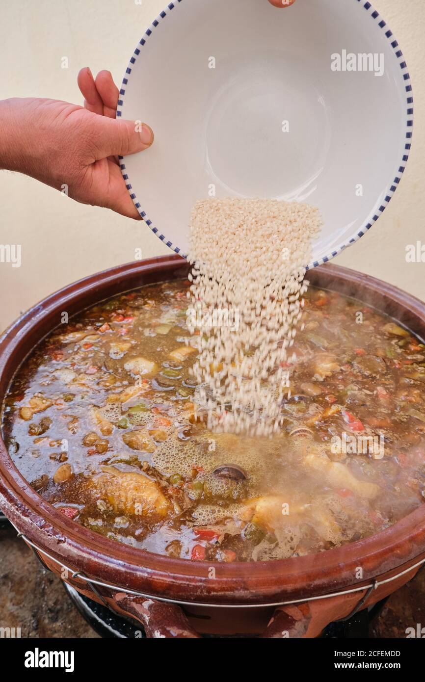 Crop mani di riso umano anonimo versando a grande argilla pentola con cottura piatto caldo di pollo verdure assortite e. Lumache di Borgogna Foto Stock