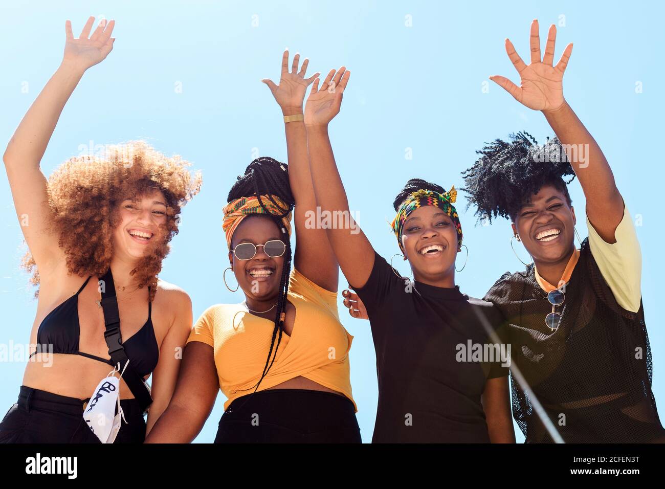 Basso angolo di compagnia di donne multietniche amichevoli che esprimono gioia con braccia sollevate sullo sfondo del cielo senza nuvole in estate e guardando la fotocamera Foto Stock