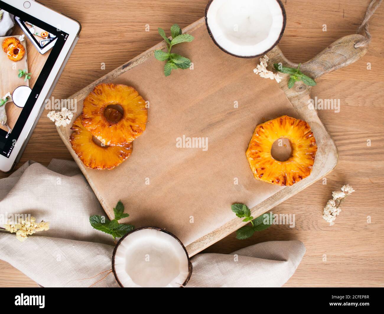 Foglie di menta fresca poste su tavola di legno vicino a fette di ananas dolce e metà di cocco gustoso su legname piano portapaziente vicino al tablet e al tovagliolo Foto Stock