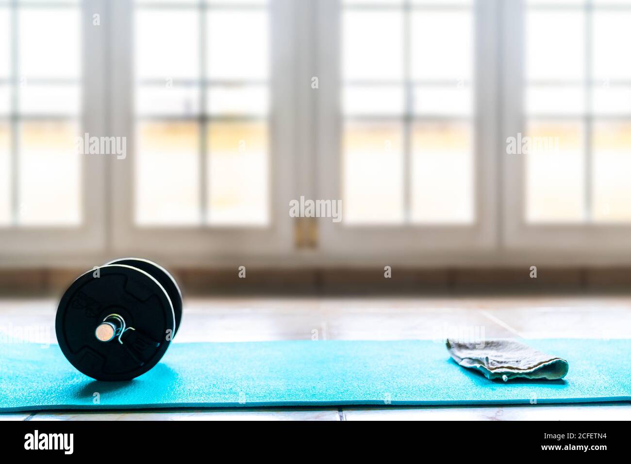 Dall'alto set sportivo per l'allenamento costituito da nero pieghevole manubri e asciugamano bianco su tappetino sportivo azzurro pavimento contro la parete della finestra sfocata in una spaziosa palestra contemporanea Foto Stock