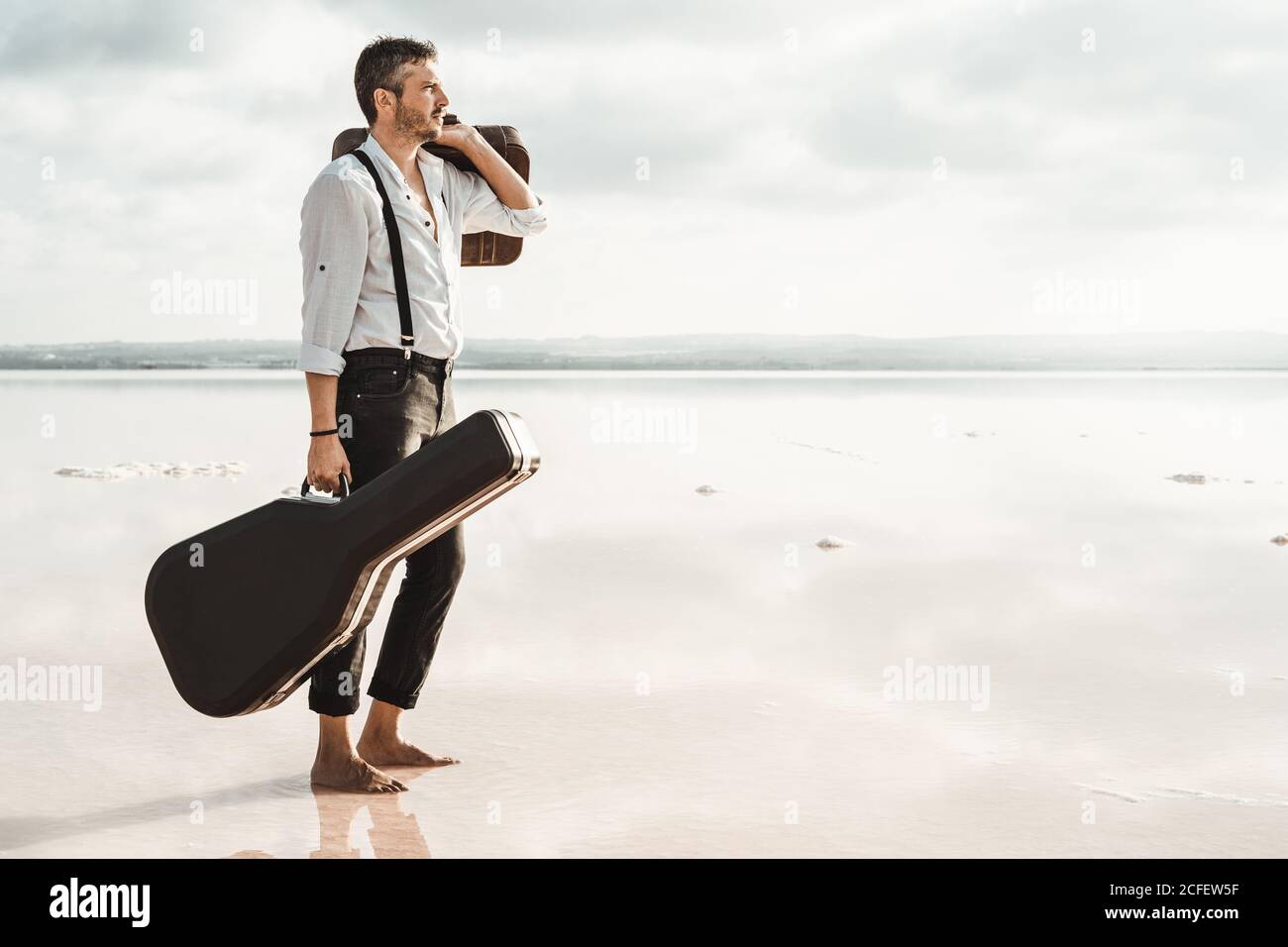 Vista laterale di un uomo serio in camicia bianca e sospetti porta la chitarra e la valigetta mentre ti trovi a piedi nudi in acqua Nuvoloso negli Stati Uniti Foto Stock