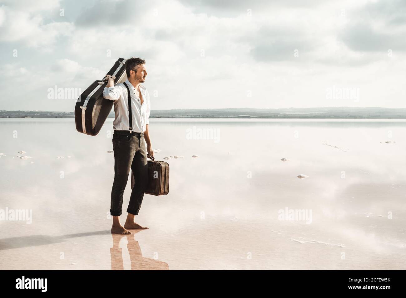 Vista laterale di un uomo serio in camicia bianca e sospetti porta la chitarra e la valigetta mentre ti trovi a piedi nudi in acqua Nuvoloso negli Stati Uniti Foto Stock