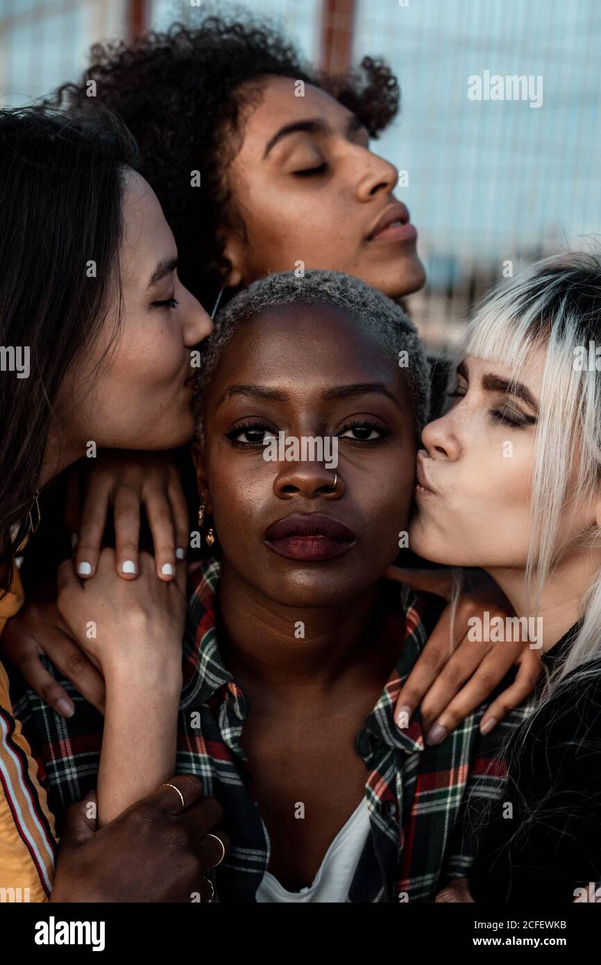 Ritratto di giovane nero a capelli corti Donna guardando la macchina fotografica con il volto serio mentre le amici femminili sostengono con abbracci e baci Foto Stock
