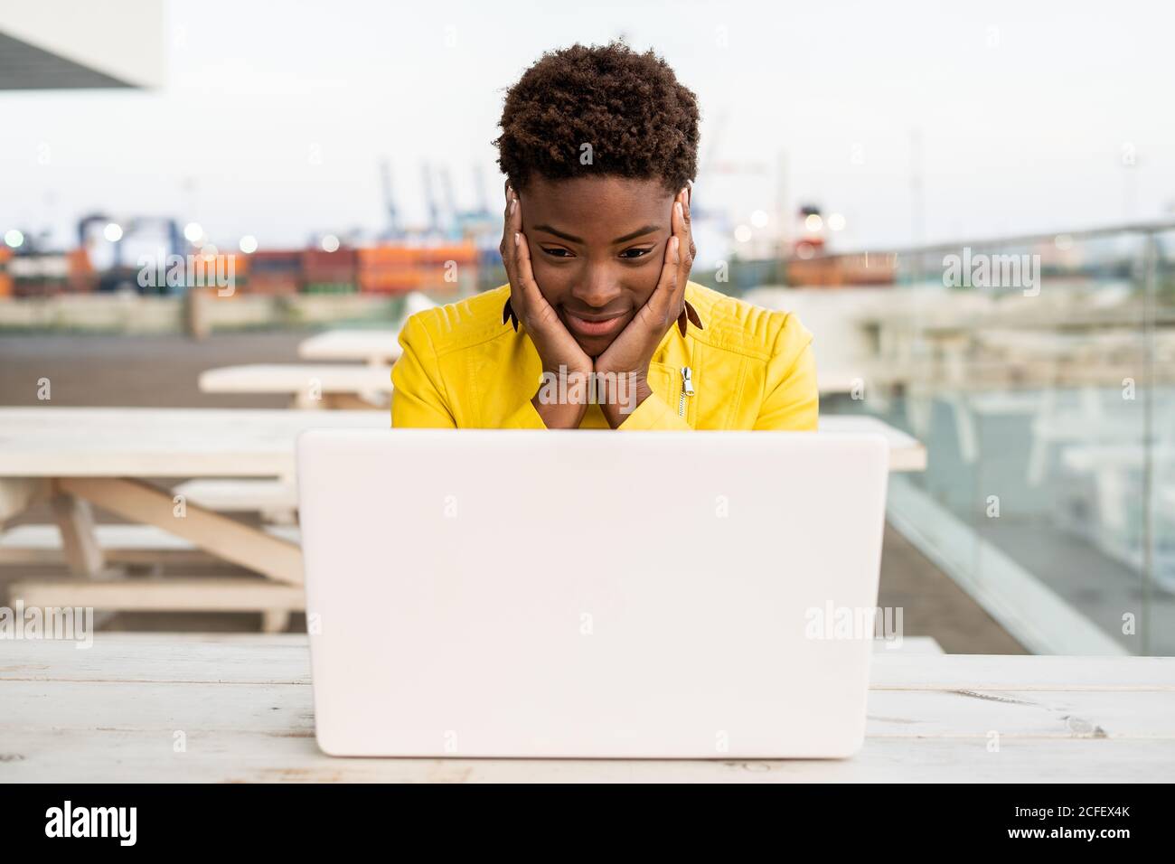 Sorpresi il volto di donna nera afro-americana in giacca gialla utilizzando un computer portatile alla scrivania di legno in città su sfondo sfocato Foto Stock