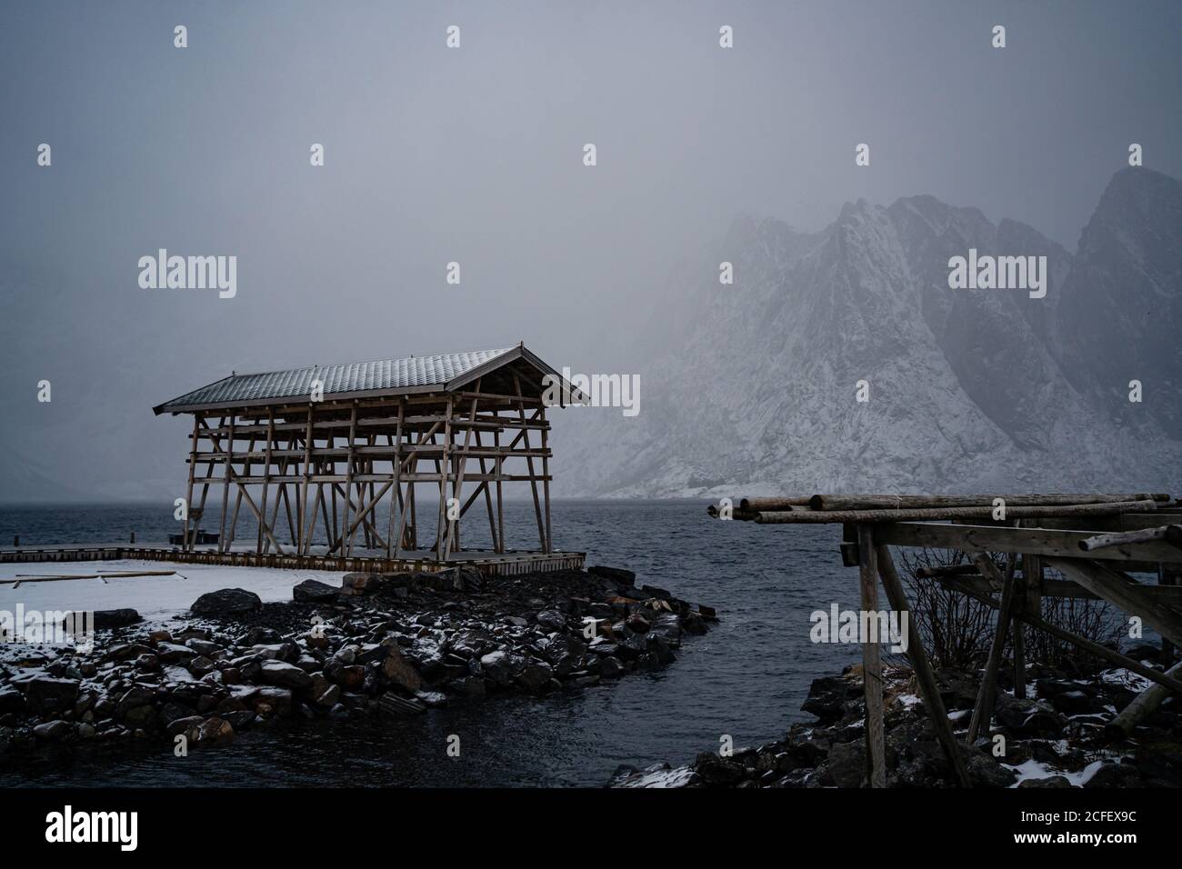 Costruzione solitaria con pilastri in legno e tetto grigio su pietra spiaggia di lavaggio da acqua tormentata contro nebbie creste di montagna in Tempo di pioggia nel porto in Norvegia Foto Stock