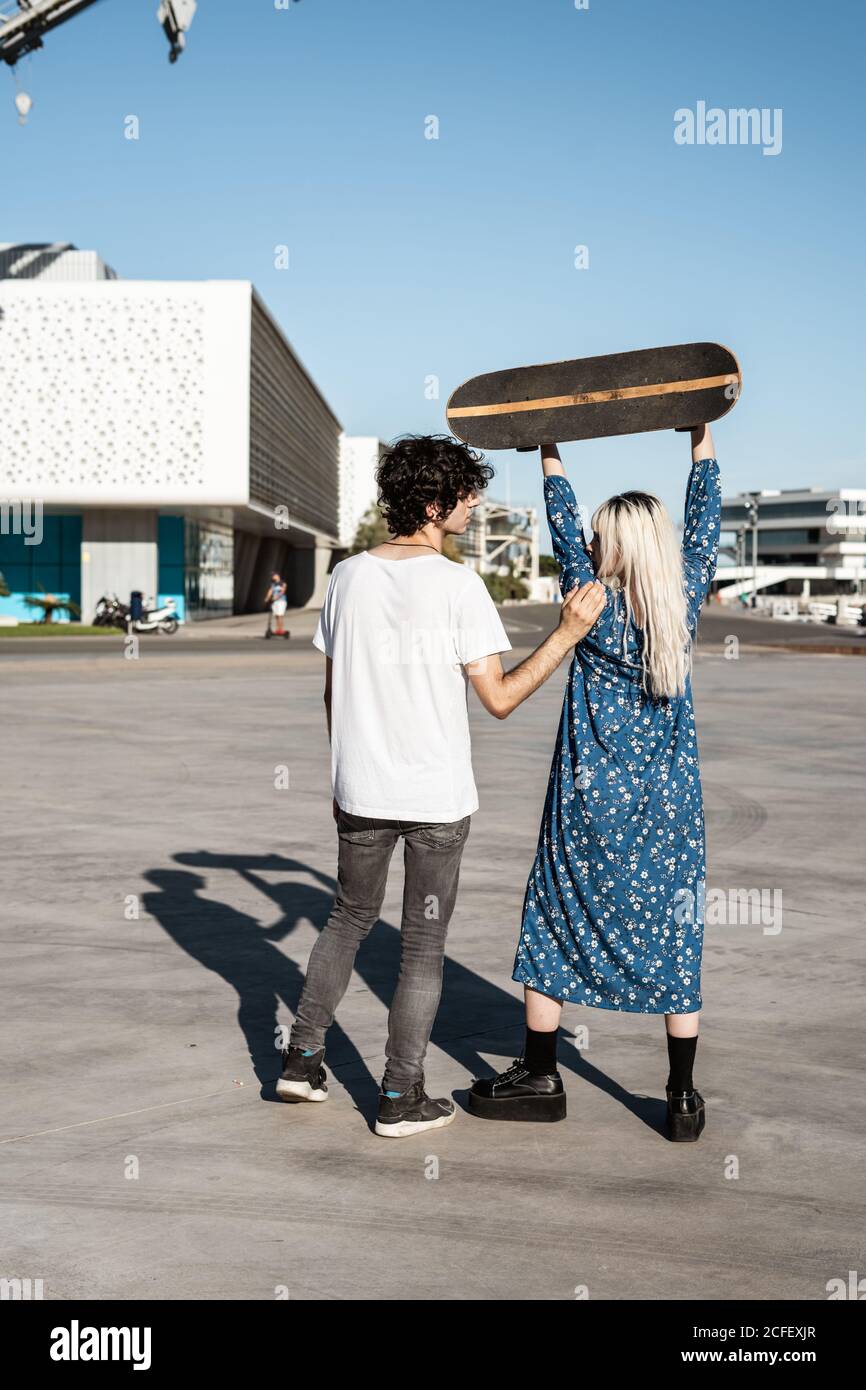 Vista posteriore di una giovane coppia alla moda irriconoscibile in piedi, mentre Woman solleva uno skateboard sulla piazza contro il cielo blu e gli edifici moderni sfocati in condizioni di vento Foto Stock