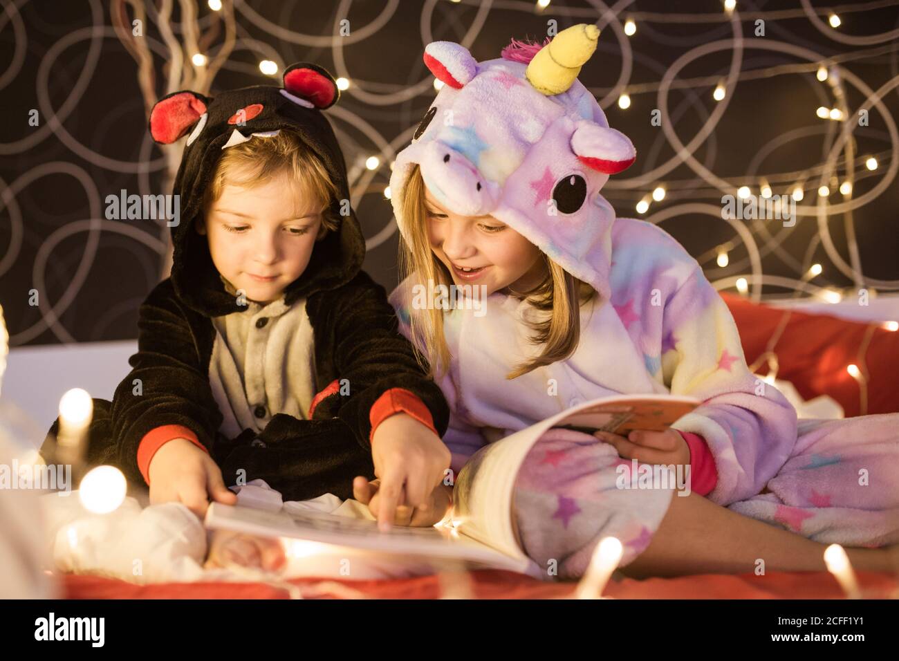 Carino piccoli fratelli in accogliente kigurumi pigiama lettura libro con Fiabe mentre si siedono insieme sul letto decorato con Natale luci Foto Stock