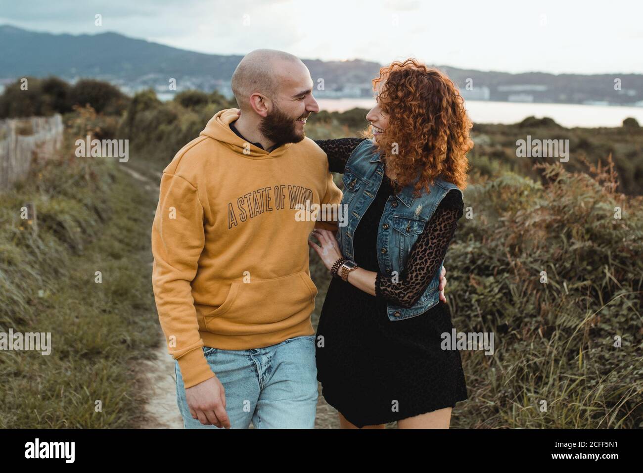 Amorevole donna tenera che tocca il mento dell'amato uomo mentre cammina insieme lungo una passerella verde fattoria sul mare con edifici di montagna e città sullo sfondo Foto Stock