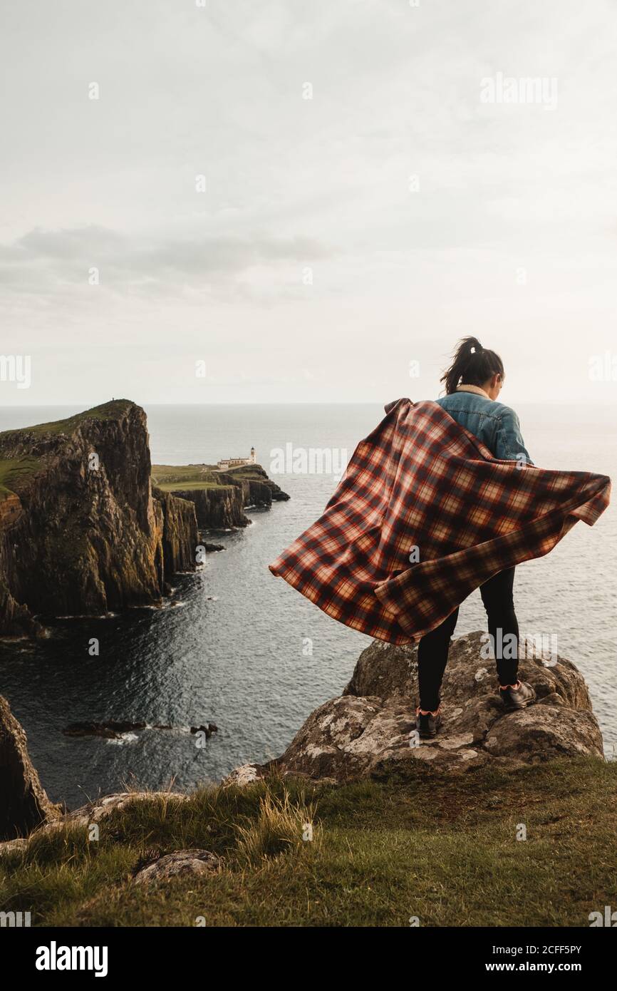 Vista posteriore di Solitary Woman con la calza a scacchi in piedi sul bordo della scogliera e guardando giù contro la falda rocciosa offuscata e la stupefacente stagione marina in una giornata ventosa in Scozia Foto Stock
