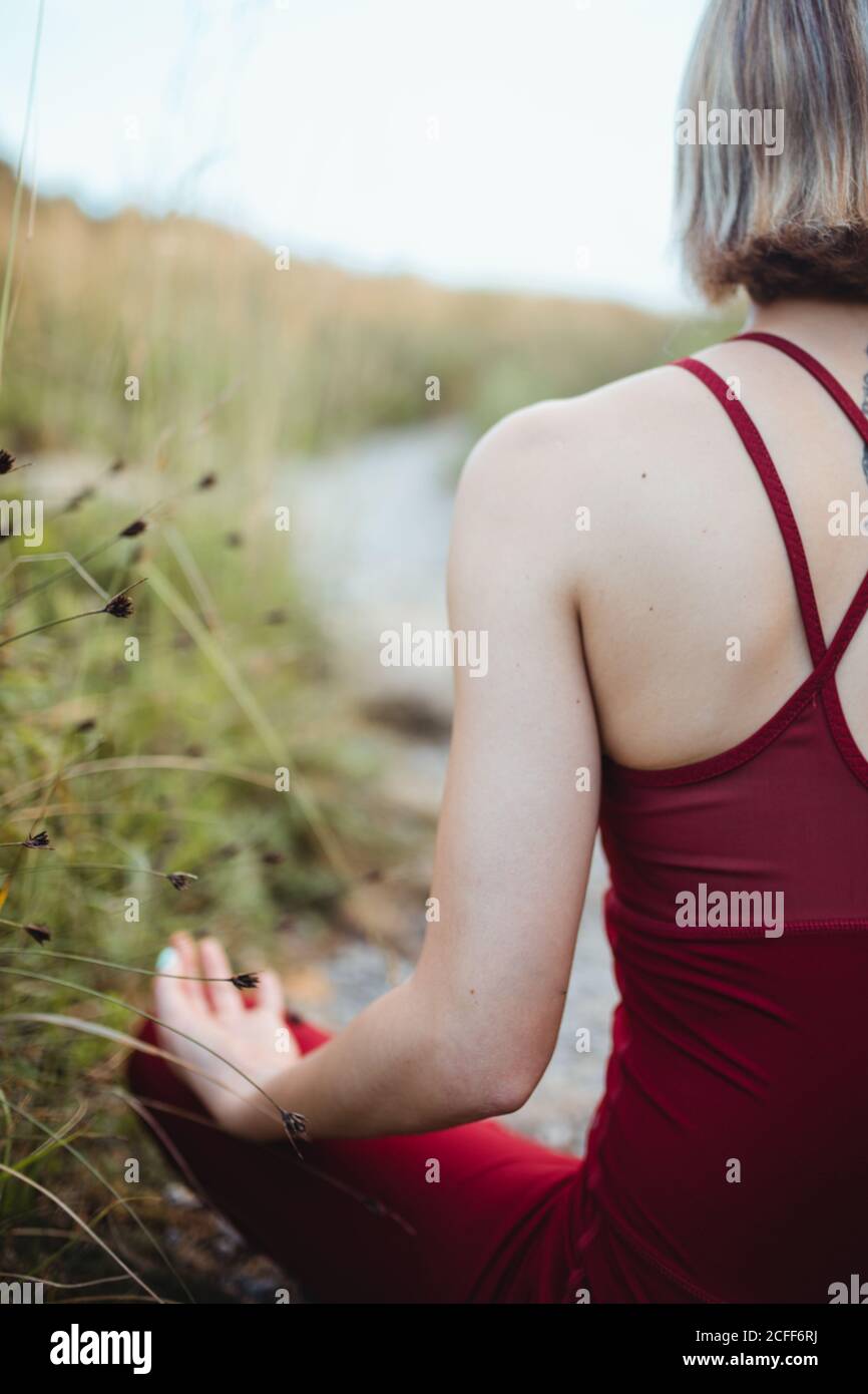 Vista posteriore di donna irriconoscibile in rosso abbigliamento sportivo allenarsi e praticare il loto posa yoga con le mani mudra mento su spiaggia rocciosa vuota Foto Stock