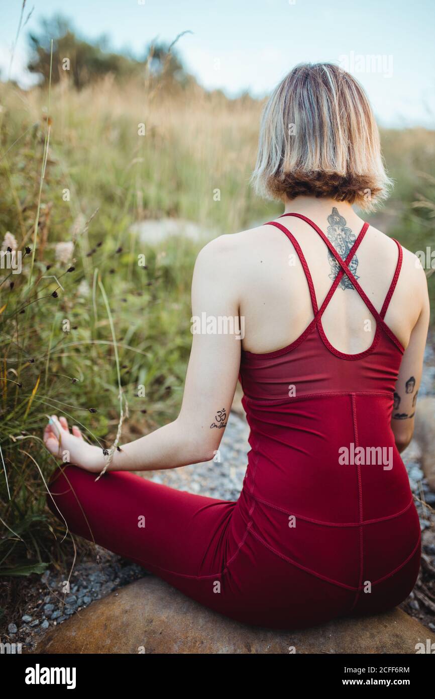 Vista posteriore di donna irriconoscibile in rosso abbigliamento sportivo allenarsi e praticare il loto posa yoga con le mani mudra mento su spiaggia rocciosa vuota Foto Stock