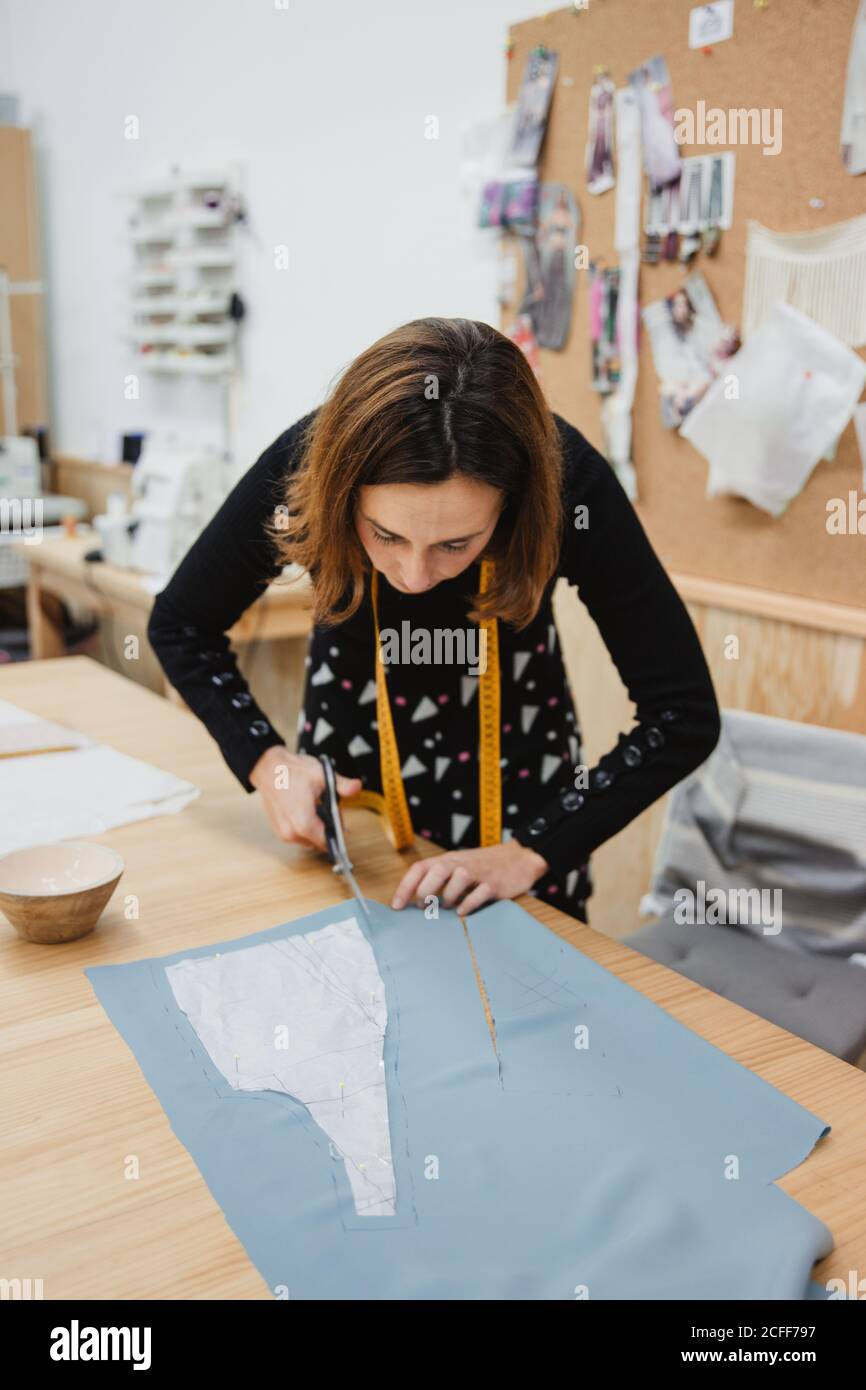 Donna che usa forbici affilate per tagliare i dettagli del capo dal tessuto seduto al tavolo in un'officina su misura Foto Stock