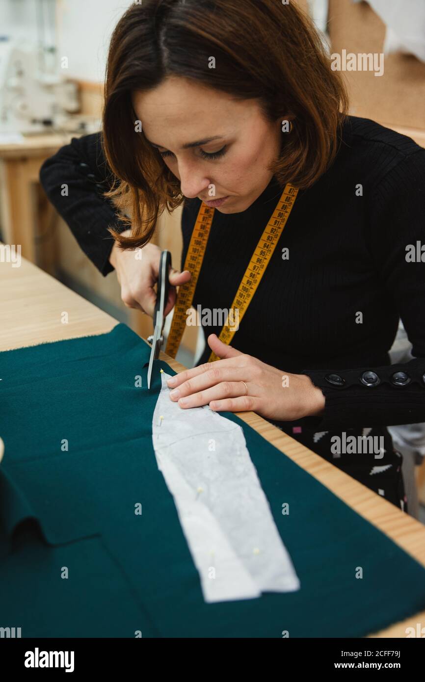 Donna irriconoscibile utilizzando forbici affilate per tagliare i dettagli del capo dal tessuto mentre si siede al tavolo in laboratorio su misura Foto Stock