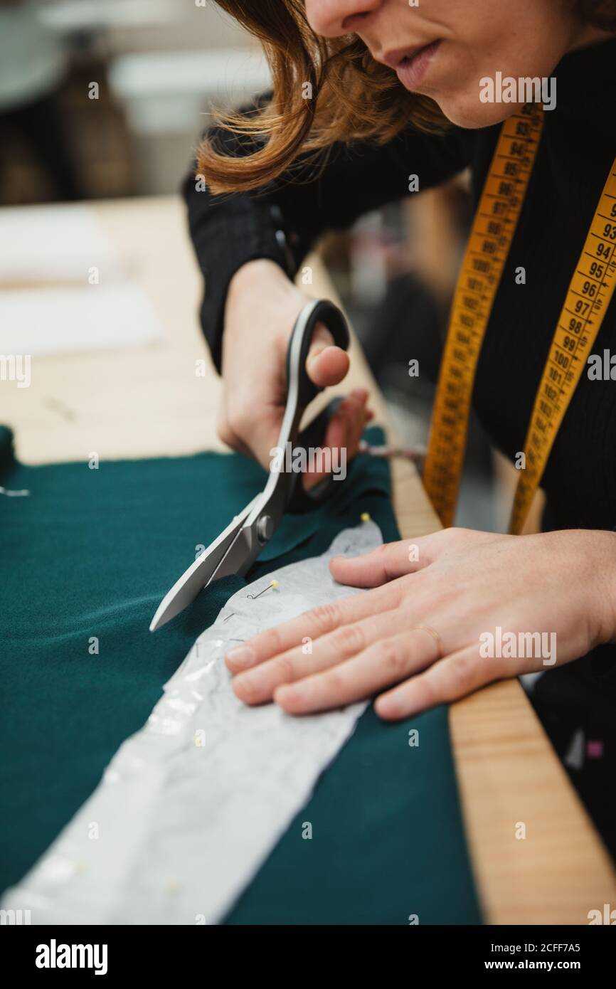 Donna irriconoscibile utilizzando forbici affilate per tagliare i dettagli del capo dal tessuto mentre si siede al tavolo in laboratorio su misura Foto Stock