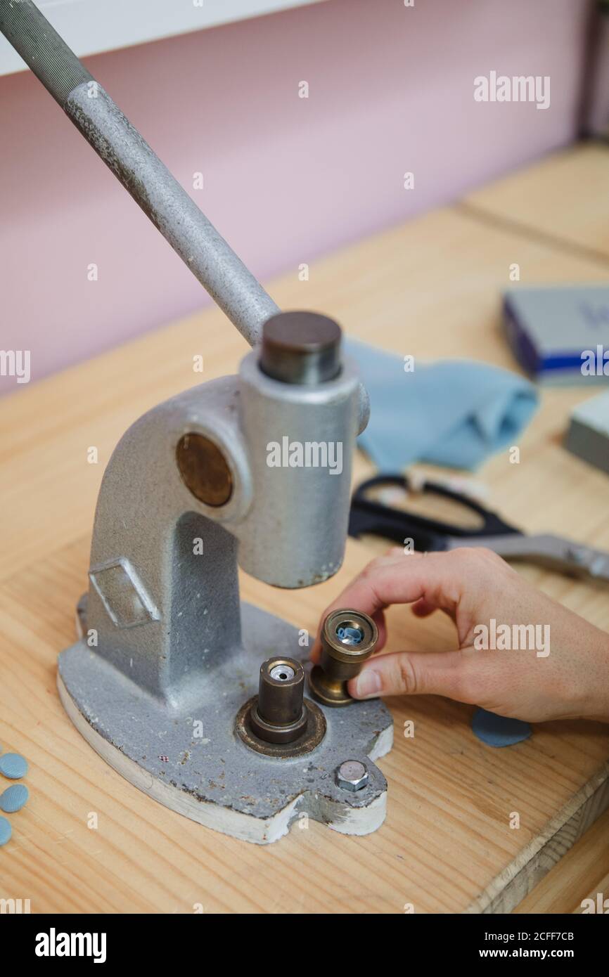 Crop persona anonima mani tirando la leva del costruttore di pulsanti su tavolo in laboratorio professionale di vestiario mentre si fa l'indumento Foto Stock