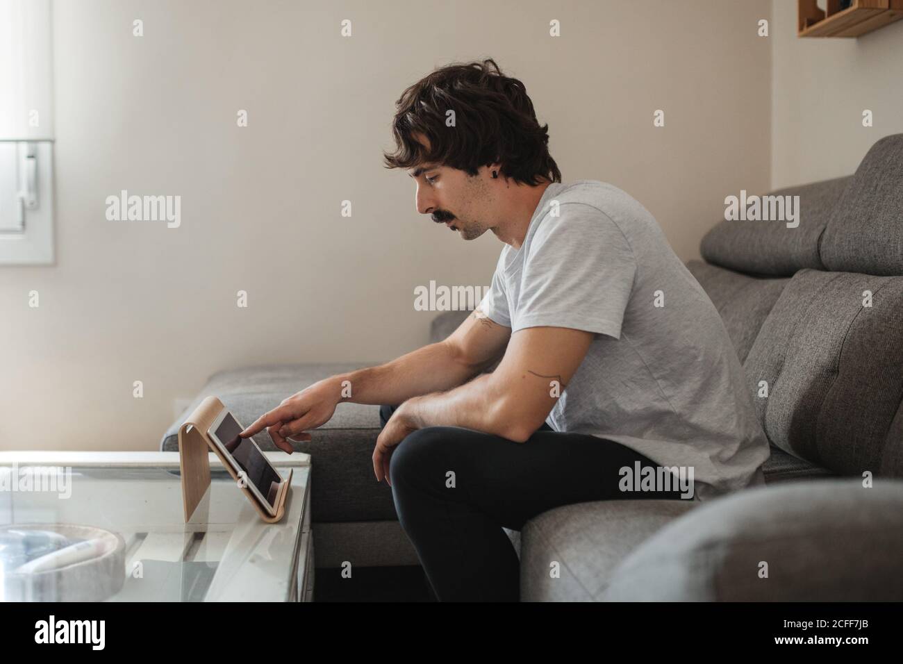 Uomo rilassato in abbigliamento casual seduto sul divano e facendo videochiama sul tablet mentre ti godi il weekend a casa Foto Stock