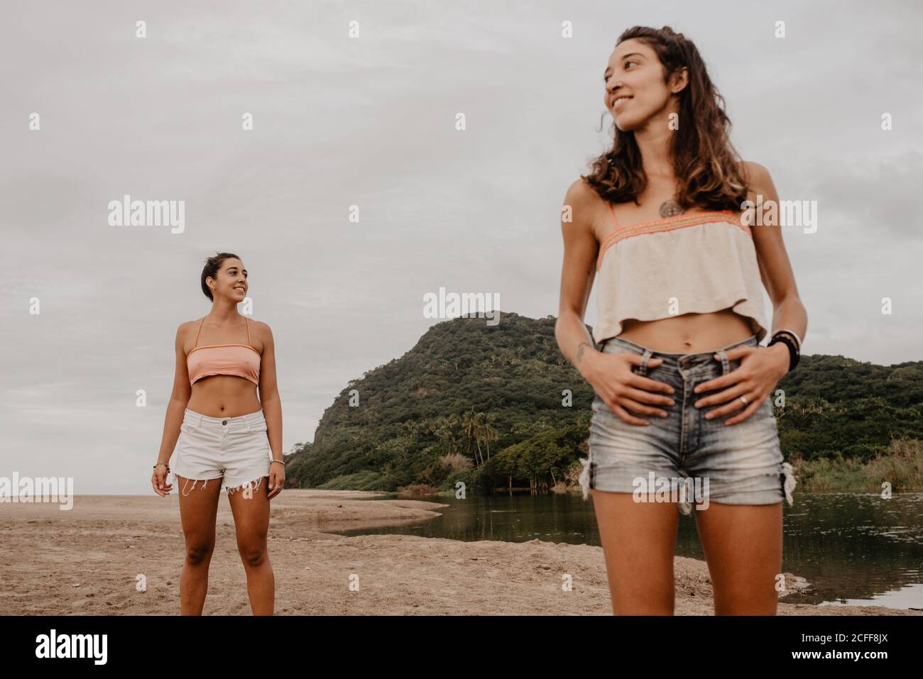 Due ragazze sottili in shorts e bras sorridenti e. guardando i lati diversi mentre si è in piedi sulla spiaggia di sabbia contro cielo grigio nuvoloso Foto Stock