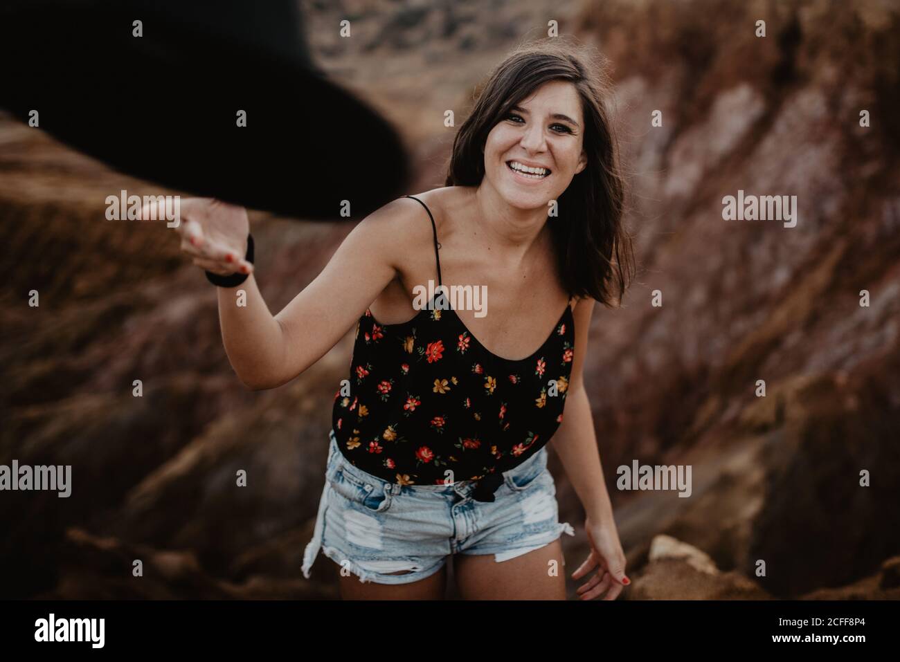 Donna elegante e sicura con camicia fiorita e pantaloncini in denim ridendo alla macchina fotografica e gettando il cappello nero mentre si levano in piedi contro collina rocciosa marrone offuscata Foto Stock