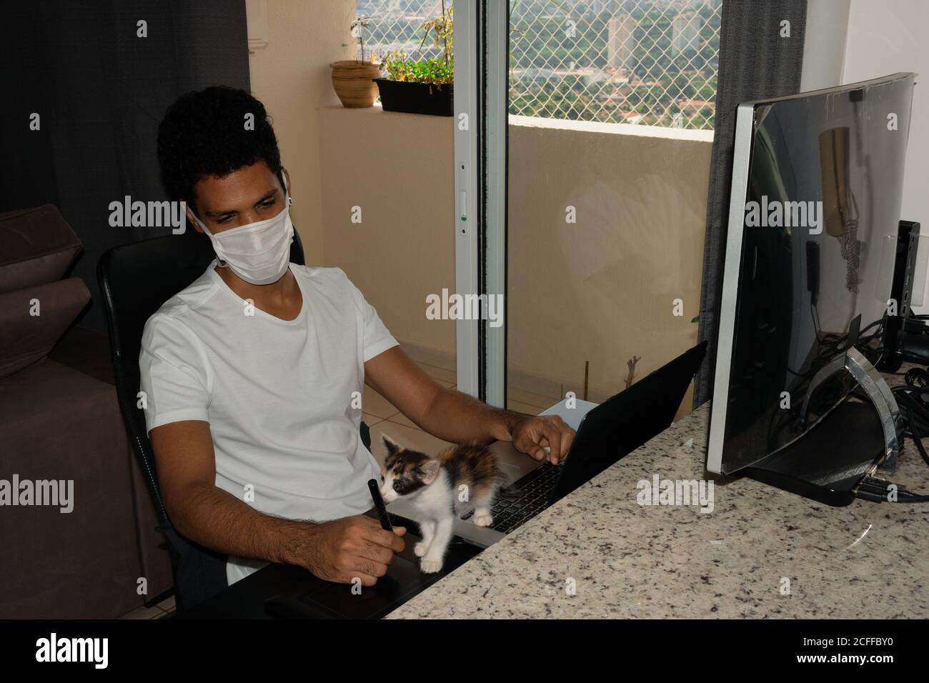 Uomo nero alla scrivania del computer con maschera pandemica con gattino. Foto Stock