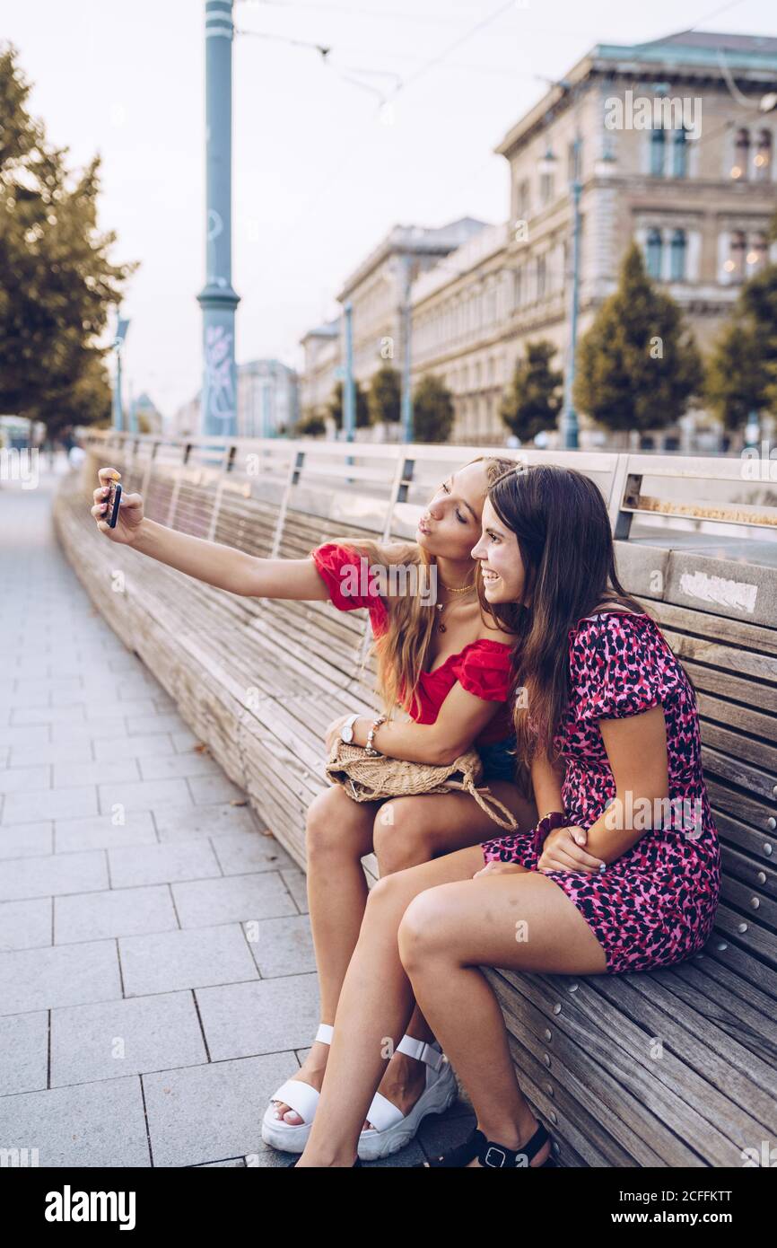 Donne sorridenti attive in un vestito colorato che prende selfie comodo posto su lunga panca di legno sullo sfondo dell'antico edificio A Budapest Foto Stock Donne sorridenti attive in un vestito colorato che prende selfie comodo posto su lunga panca di legno sullo sfondo dell'antico edificio A Budapest Foto Stock
