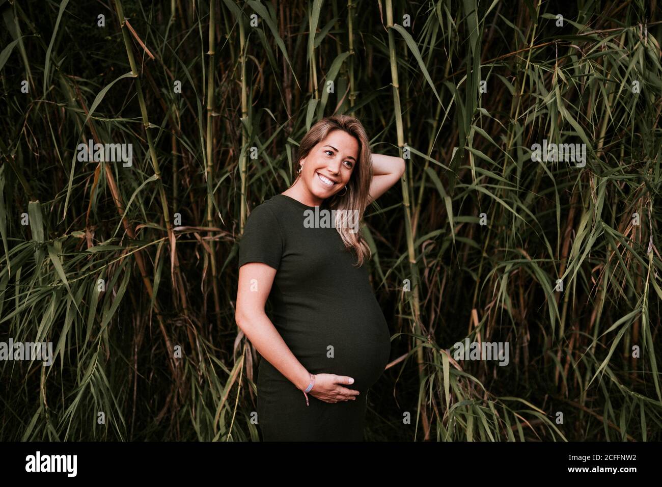 Donna incinta felice in vestito casual che stropicca il ventre mentre in piedi sulla parete verde degli alberi in giorno di estate soleggiato con cielo blu Foto Stock