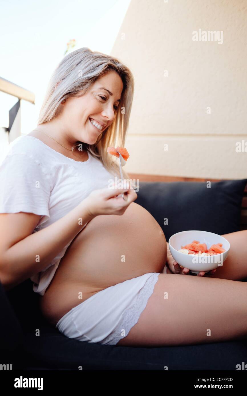 Donna incinta che indossa una maglietta bianca che mangia banane affettate e anguria dalla ciotola con forchetta mentre si siede sul divano scuro Foto Stock