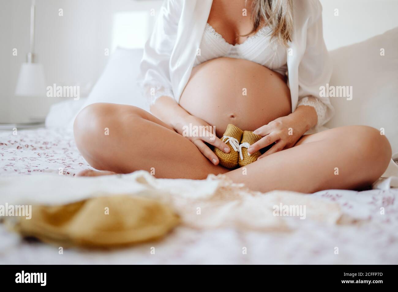Crop femmina incinta senza volto vestita in blusa bianca aperta e. bra mostrando abiti per non nato mentre si siede a letto con gambe incrociate Foto Stock